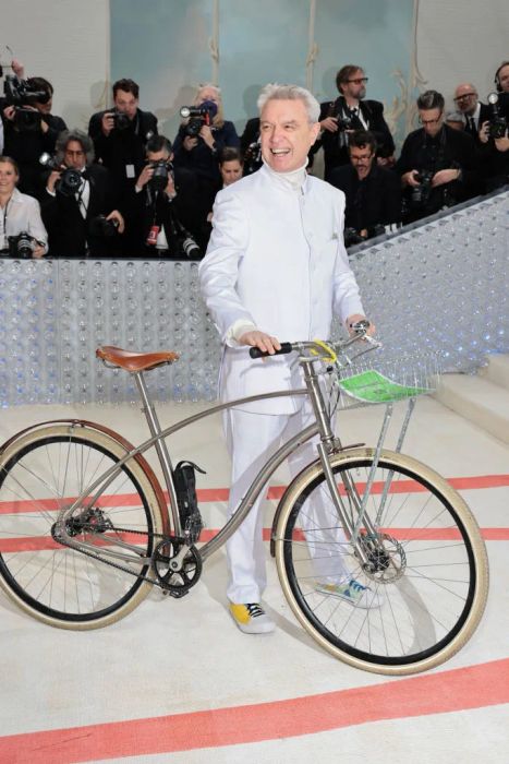David stands on the red carpet in a sleek white suit, holding a bicycle, as photographers crowd around him