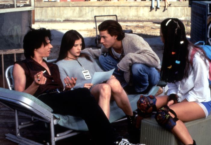 Four individuals are lounging outdoors, casually reading a newspaper and chatting. They are dressed in relaxed, youthful outfits and accessories