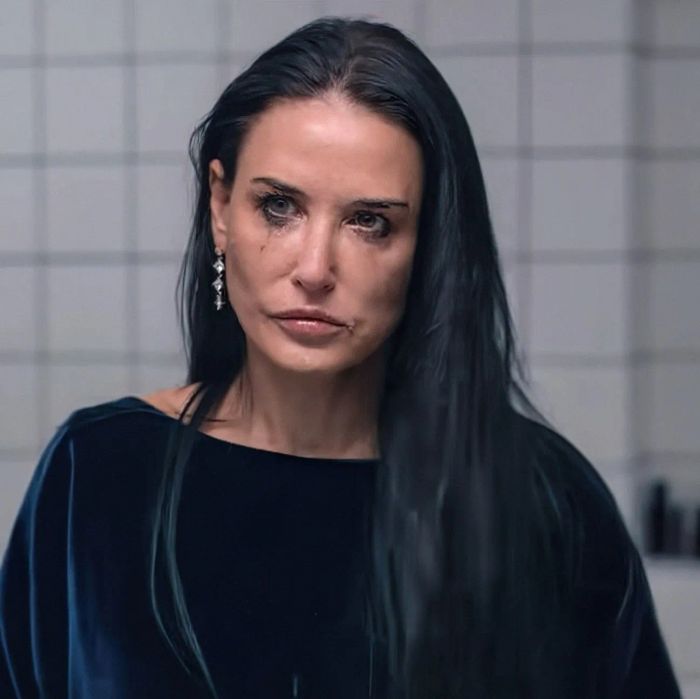 Demi, with her long dark hair, gazes at her reflection in a tiled bathroom mirror, dressed in a dark top, exuding a contemplative mood.