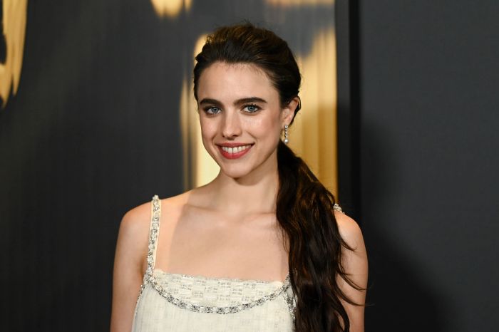 Margaret smiles while wearing a sleeveless, elegant dress with intricate beaded details and long earrings at a red carpet event.