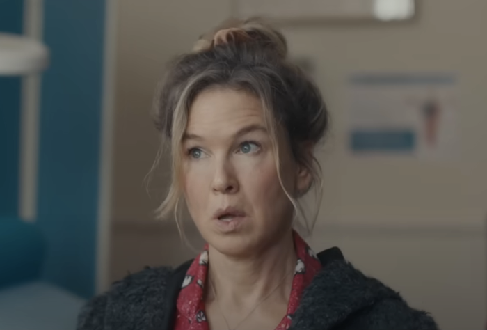Renee as Bridget Jones, looking surprised while wearing a patterned red top and a dark textured outer jacket, inside a room.
