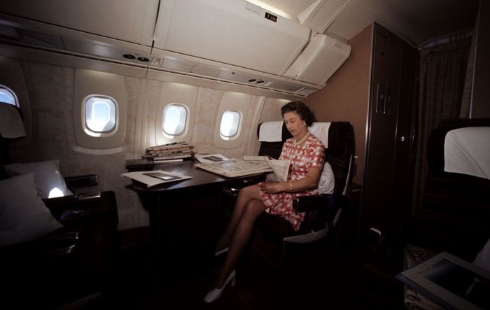 A person in a checkered dress sits in an airplane cabin, reading a newspaper with papers resting on a table beside them.