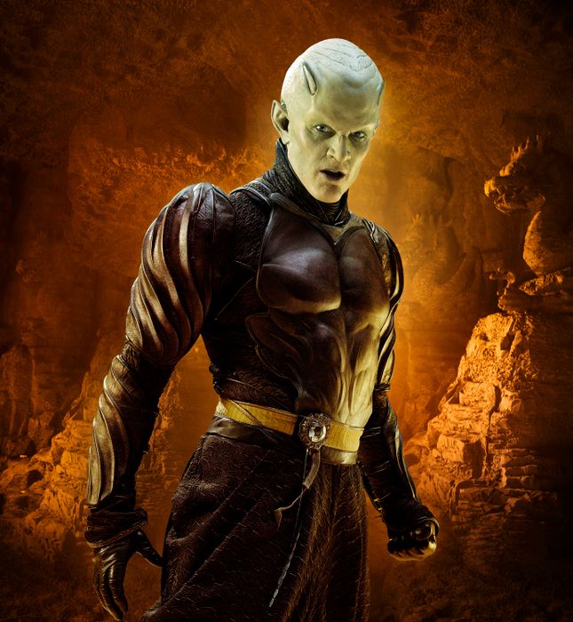 James, fully made up as an alien and dressed in intricate leather armor, stands in a vast cavern with rocky formations surrounding him.