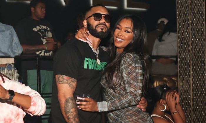 A man dressed in casual streetwear and a woman wearing a patterned jumpsuit smile together at an event.