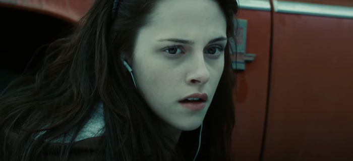 Kristen, with long hair and wearing earphones, stares intently at something off-camera, looking concerned, with a vehicle visible in the background.