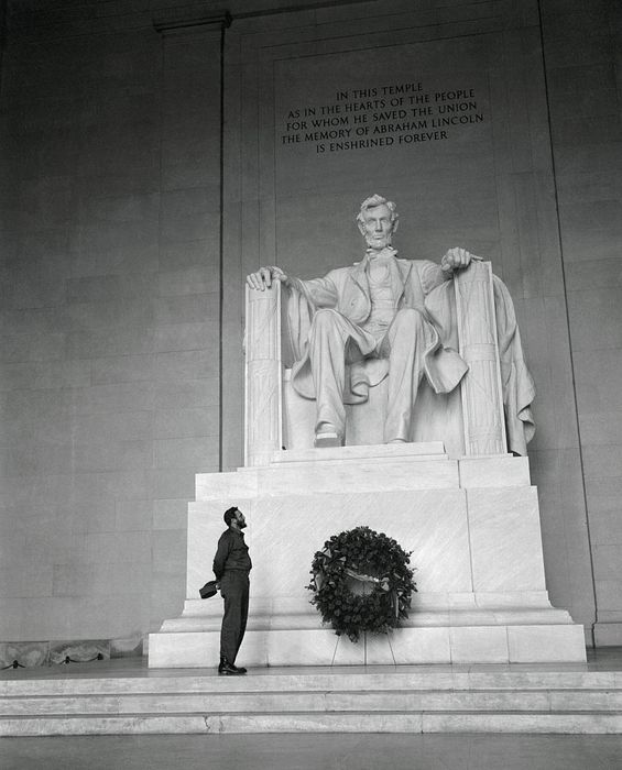 A person stands in front of the Lincoln Memorial, gazing up at the large seated statue of Abraham Lincoln, with a wreath placed at its base.
