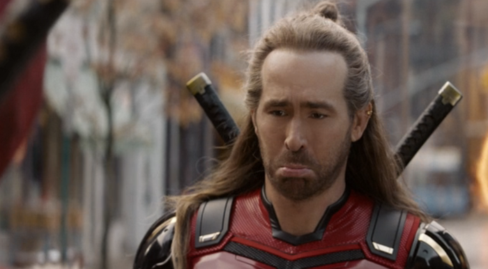 A person dressed as a warrior with a sorrowful expression, wearing armor and two swords on their back, standing in a street setting.