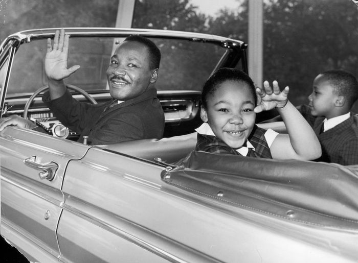 Martin Luther King Jr. waves cheerfully from a convertible car with his daughter Yolanda and son Martin Luther King III beside him.