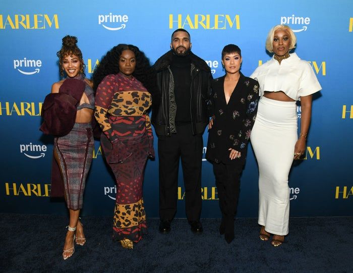 A group of celebrities poses on a blue carpet, dressed in a mix of trendy and eclectic outfits, such as patterned dresses, a leather jacket, and a stylish two-piece set.