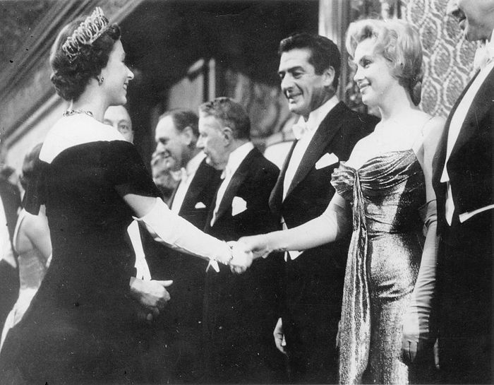 Queen Elizabeth II and Marilyn Monroe exchange a handshake while Laurence Olivier observes, all dressed in formal attire at a historic event.