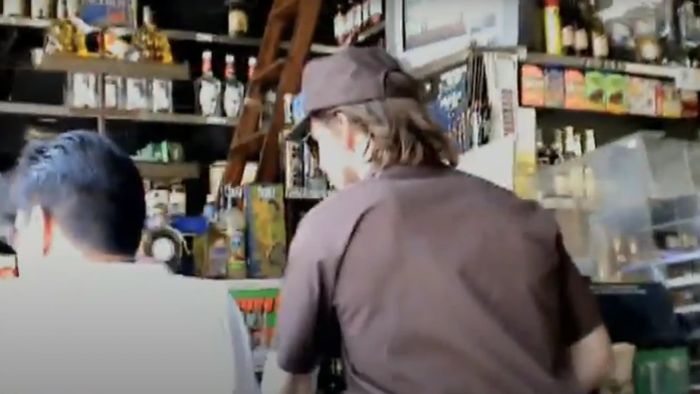 In a shop, two people stand at a counter surrounded by various products on shelves. Tom Cruise, wearing a cap and uniform, appears casually involved in the scene.