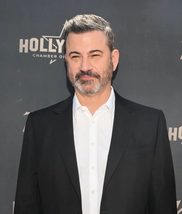 A person dressed in a suit at a Hollywood event, standing before a logo backdrop