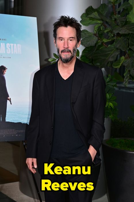A person in a black suit and shirt, standing beside a movie poster at an event, with a plant in the background