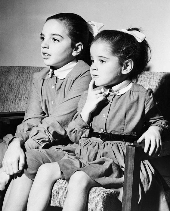 Two young girls, one thoughtfully resting her finger on her chin, sit on a couch wearing matching long-sleeved dresses.