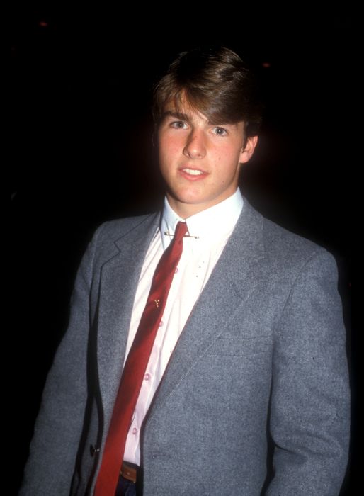 A person dressed in a formal suit, including a suit jacket, white shirt, and red tie, posing against a dark background
