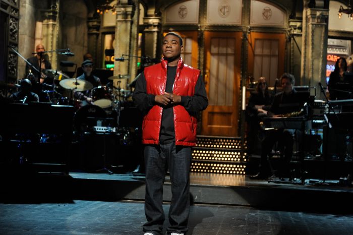 Tracy, dressed in a vest and jeans, stands on stage with musicians performing in the background.