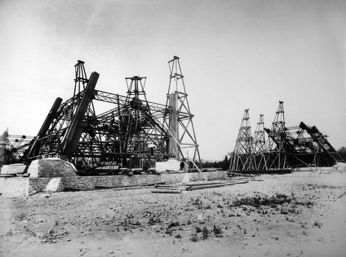 A historic black-and-white image showing the Eiffel Tower in the early phases of construction, with the iron framework partially erected on the ground.
