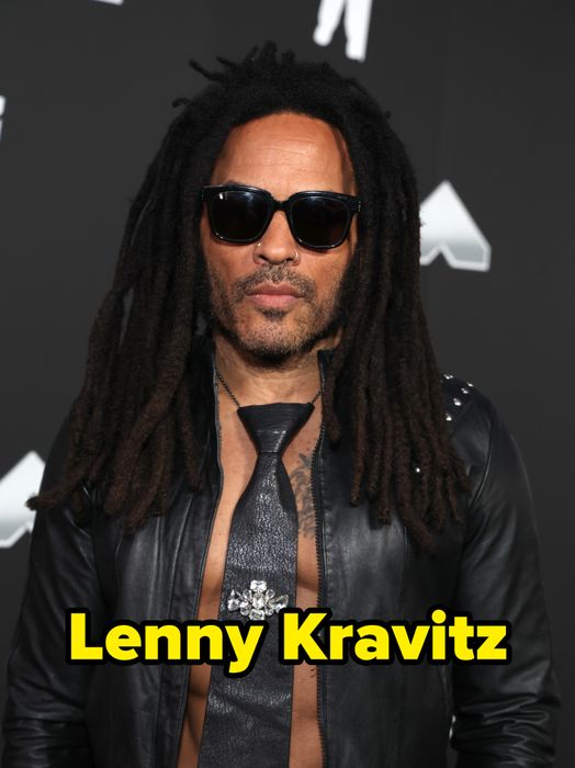 A person with locs, wearing sunglasses, a black leather jacket, and a textured tie adorned with a brooch, standing on a red carpet