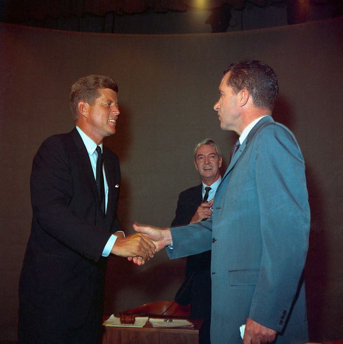 John F. Kennedy and Richard Nixon shaking hands during the 1960 presidential debate, with a man in a suit smiling in the background.