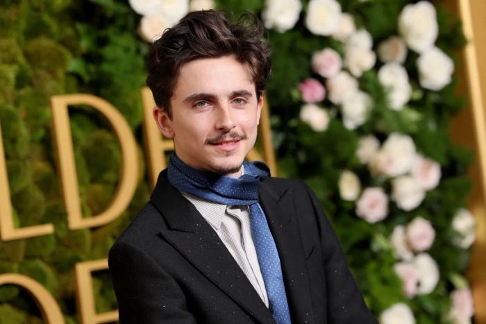 A man on the red carpet in a dark suit, striped shirt, and blue tie, standing before a floral backdrop