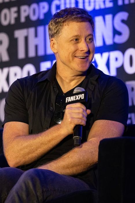 Alan beams with a smile while seated on stage at a Fan Expo event, microphone in hand, with event branding in the background.