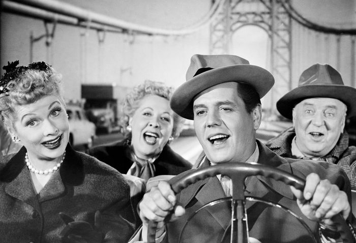 A classic black-and-white photo of four actors joyfully riding in a convertible, with a bridge in the background. They sport 1950s-style hats and coats.