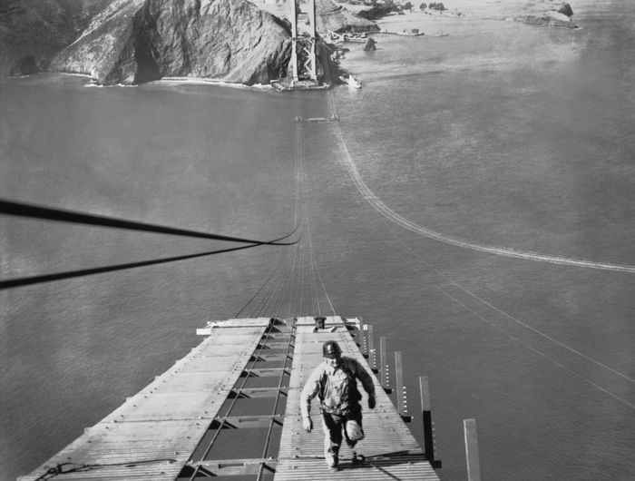 A construction worker carefully walks along the unfinished Golden Gate Bridge framework, with the scenic San Francisco Bay and distant hills visible in the background.