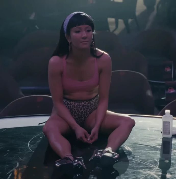 Constance is seated on a mirrored stage, wearing a crop top, leopard print shorts, high heels, hoop earrings, and a headband.