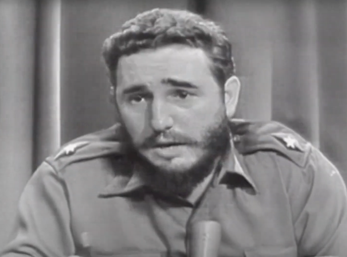 A bearded man dressed in a military uniform, sitting in front of a microphone, participating in a broadcast interview.