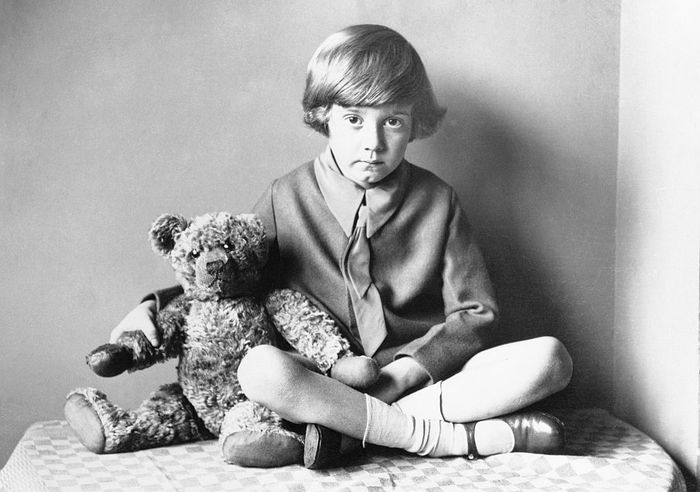 A young child sits cross-legged with a teddy bear, wearing a collared sweater and shorts, gazing directly at the camera in a vintage setting.