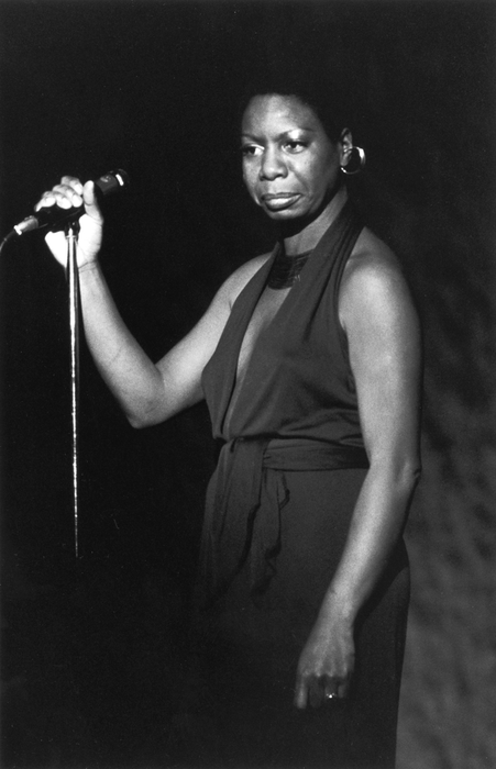 A performer stands confidently on stage with a microphone in hand, wearing a sleeveless, high-neck dress, radiating elegance and strength