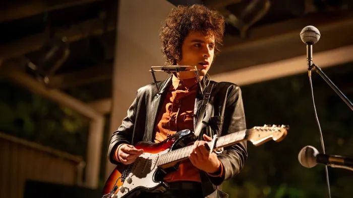 Timothée Chalamet takes the stage with a guitar and harmonica holder, dressed in a vintage-inspired jacket and shirt, performing at an outdoor venue