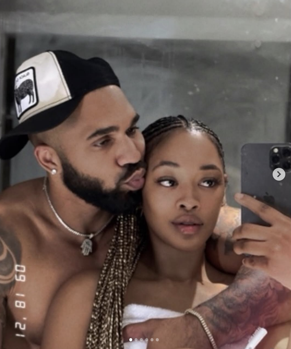 Two people take a playful mirror selfie, one with a cap and beard, kissing the other's cheek. They appear dressed casually and happily together.