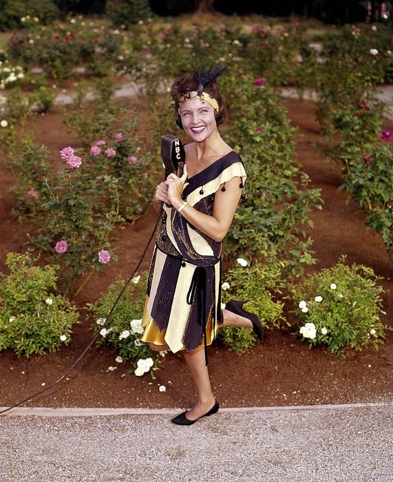 A woman joyfully poses in a vintage flapper-style dress in a garden, holding a microphone.
