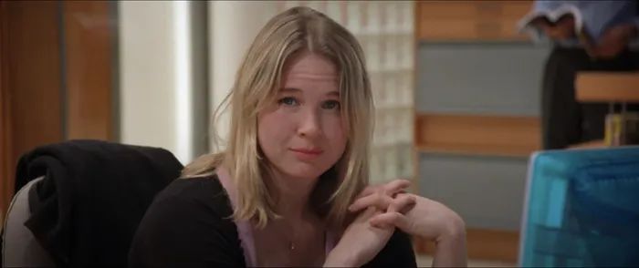 Renée sitting at a desk, dressed casually, gazing into the camera with a neutral expression. The backdrop features a typical office environment.