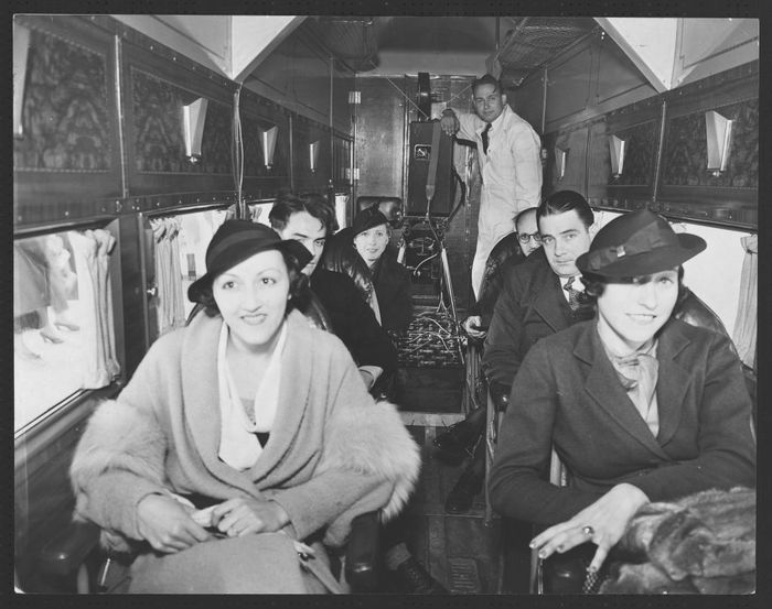 Passengers seated inside a vintage airplane cabin, dressed in early 20th-century formal attire, complete with hats and coats.