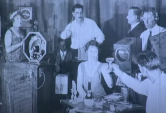 A vintage photo showing people raising glasses at a gathering, with an old-fashioned camera visible in the scene.