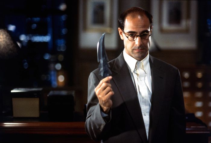 Stanley, dressed in a suit, stands inside, holding a large, curved knife and gazing at it in contemplation.