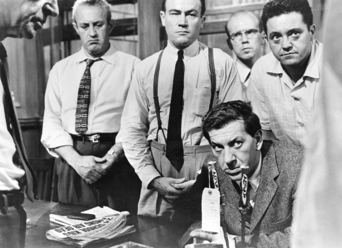 A group of men in a tense moment from a classic film, one sitting while others stand around him, all wearing formal 1950s-era clothing