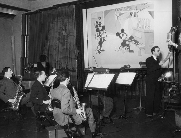 Walt Disney conducts a band of musicians recording music for an animated Mickey Mouse cartoon. The musicians play saxophones, while Disney gestures toward the screen.