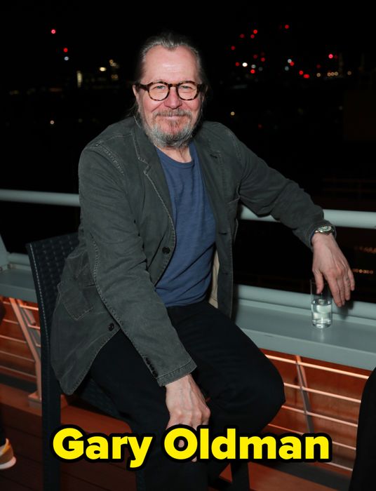 A person seated on a patio at night, wearing glasses, a jacket, and a t-shirt, with the glow of city lights in the distance