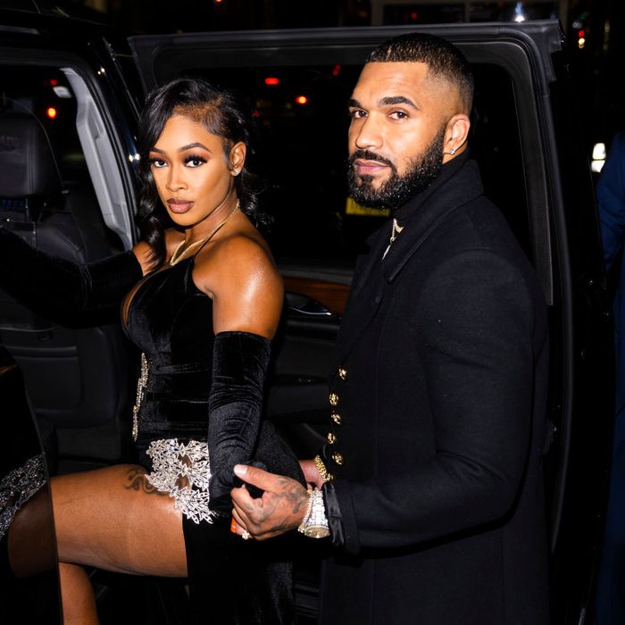 Two people step out of a car. The woman is dressed in a black velvet evening gown with silver accents, while the man wears a black coat adorned with gold buttons.