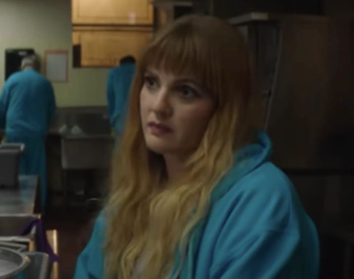 In a kitchen setting, Drew, with long hair and dressed in a casual hoodie, appears pensive while others dressed similarly work in the background.
