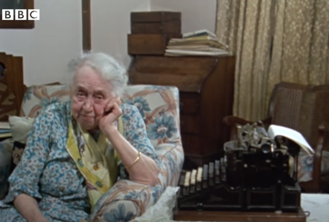 An elderly woman, sitting thoughtfully on a floral-patterned couch beside an old typewriter, with the BBC logo visible in the corner of the screen.