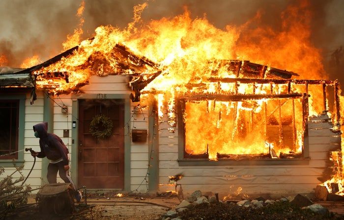 A person dousing a burning house with water as large flames and thick smoke billow from the structure.