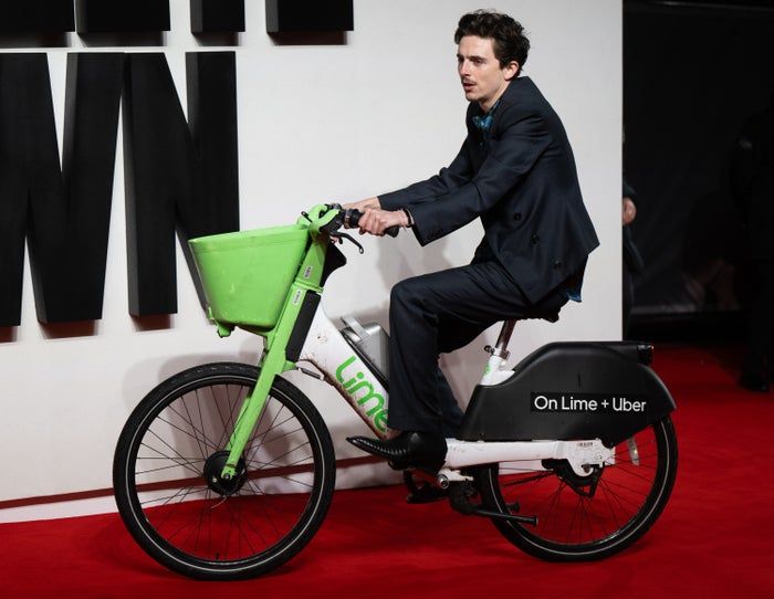 A man, dressed in a sharp suit, rides a Lime electric bike down the red carpet at an event, with large text visible in the background.