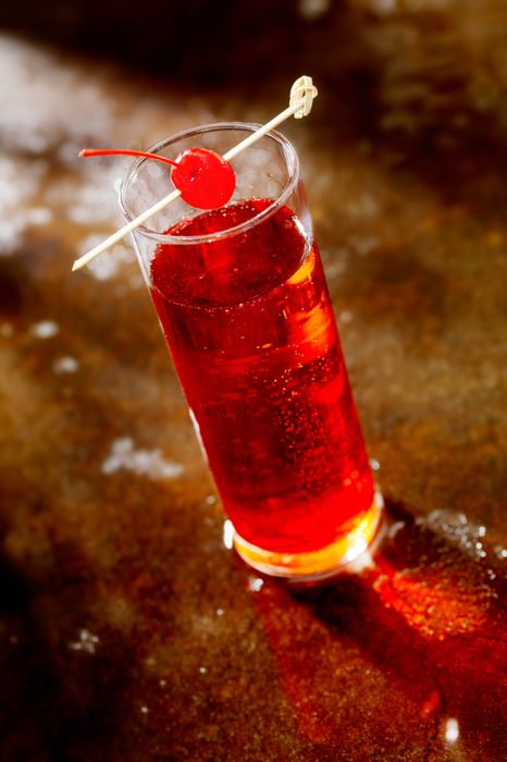 A tall glass of a fizzy beverage topped with a red cherry and a decorative skewer, placed on a table