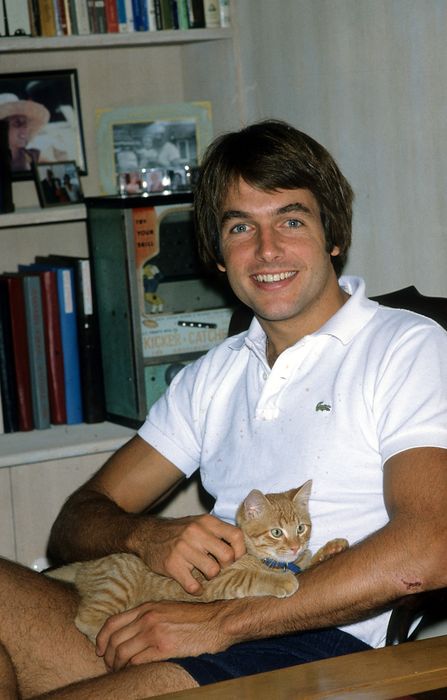 A man sits in a chair, grinning while holding a small tabby cat. Behind him, there are books and framed photographs on a shelf