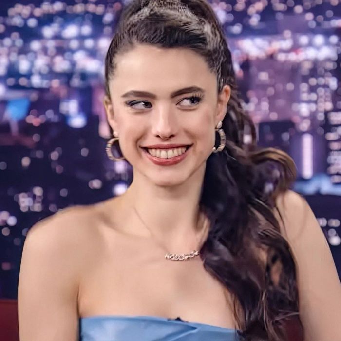 Margaret, in a strapless dress, smiles during a TV interview set against a city skyline backdrop.