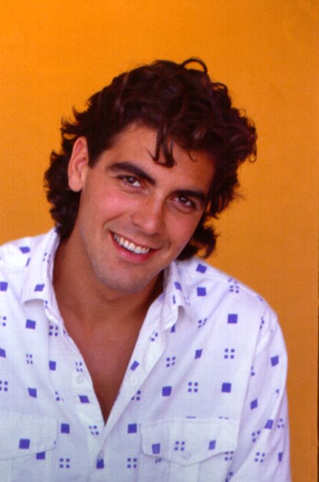 A person smiling at the camera, wearing a button-up shirt with a geometric pattern. The background is a solid color